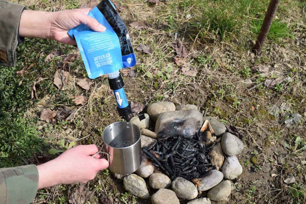 Using Sawyer Mini to filter water for drinking, pouring filtered water into a camping cup with a background of a campfire that has been put out in the woods on a sunny day