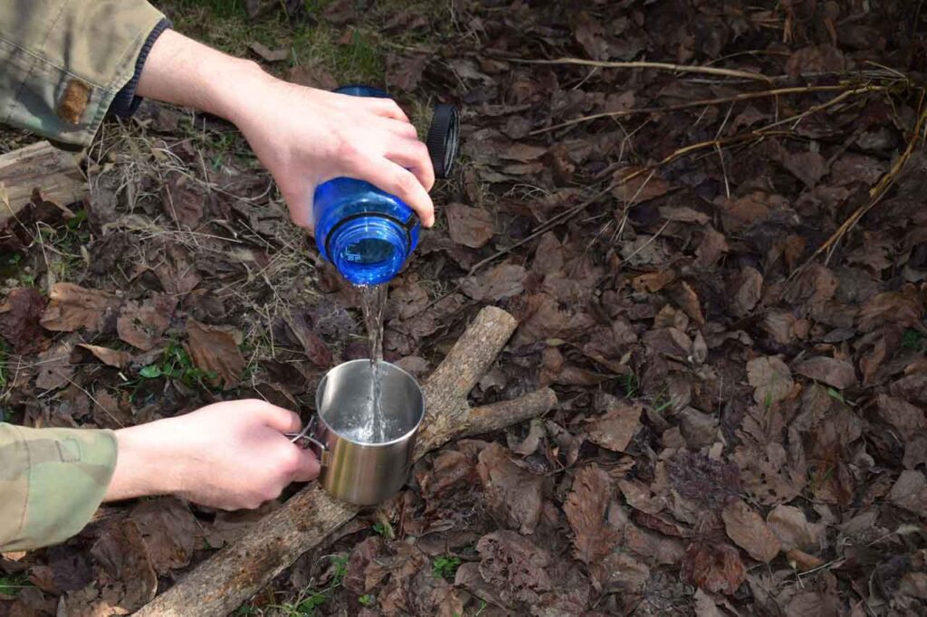 Pouring filtered water into a stainless steel cup in the woods surrounded by dry leaves