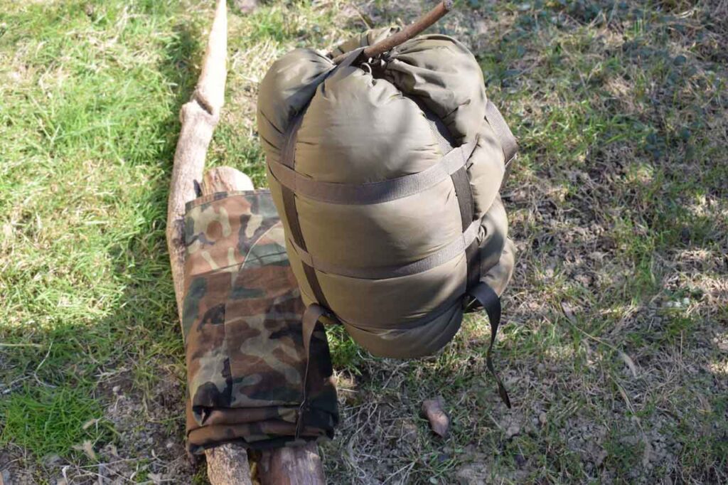 The Best Survival Shelter. Sleeping bag in a compression sack hanging on a twig with camo tarp folded on some wood on the ground surounded by green and dry grass on a sunny day