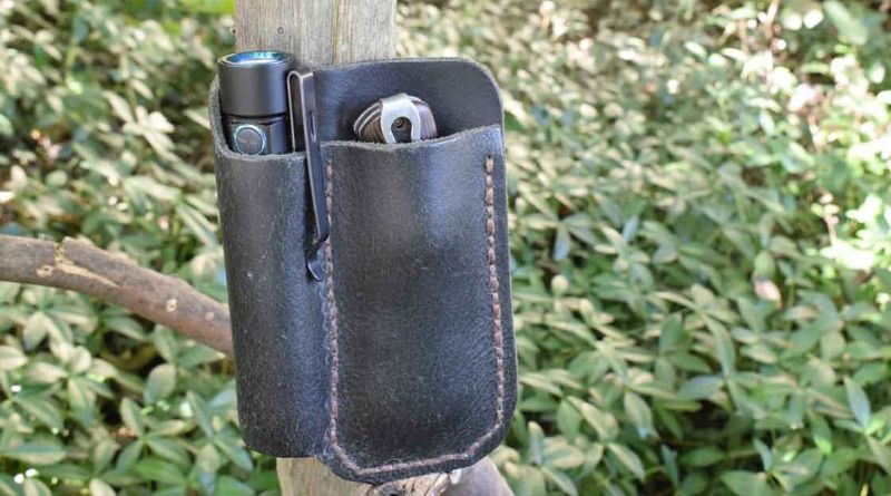 An Image Of A Home crafted Dark Brown Leather Organizer With A Survival Flashlight On One Side And A Folding Buck Knife On The Other Side With Greenery as a Backdrop